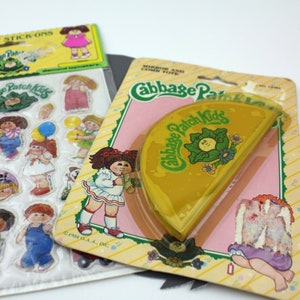 Puede incluir: Una hoja de pegatinas de Cabbage Patch Kids con una variedad de pegatinas que muestran el logotipo de Cabbage Patch Kids, un sol sonriente y diferentes muñecas Cabbage Patch Kids. La hoja de pegatinas está empaquetada con un espejo y un estuche para peine de plástico amarillo.