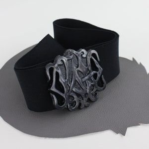 May include: Black elastic belt with a large, ornate, silver-toned buckle. The buckle features a stylized design with intricate details.