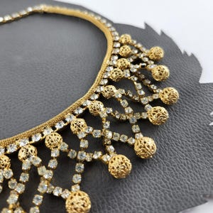 Puede incluir: Un collar dorado con una fina cadena de malla y un elaborado diseño de colgante. El collar presenta cristales transparentes brillantes y esferas de filigrana decorativas, creando una apariencia glamurosa y ornamentada.