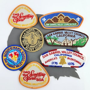 May include: A collection of seven embroidered scout patches from the San Gabriel Valley Council. The patches feature various designs, including a bell, a church, mountains, and a scout holding a flag. The patches are in various colors, including blue, red, white, yellow, and green.