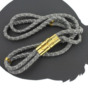 Könnte beinhalten: Ein graues geflochtenes Seil-Boloneckband mit goldfarbenen Metallspitzen. Das Seil ist in einem einfachen Muster geflochten und die Spitzen sind glatt und glänzend.