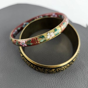 May include: Two decorative bangles. One is a dark red with floral designs in pink, white, and green. The other is gold with black floral patterns. Both are circular and appear to be made of metal.