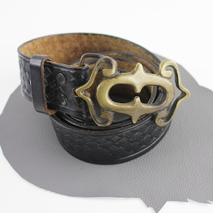 Puede incluir: Un cinturón de cuero negro con una hebilla de latón decorativa que presenta una letra C estilizada. El cinturón tiene un patrón tejido y está usado.