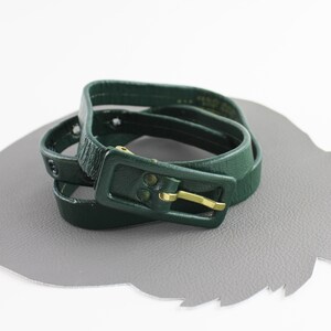 May include: A dark green leather belt with a gold buckle. The belt is wrapped around itself and has a few holes punched into it.