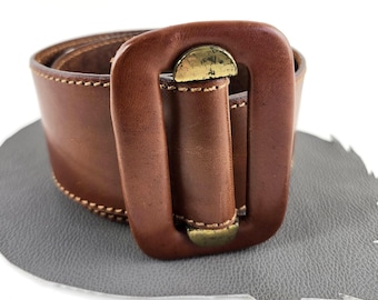 May include: A wide brown leather belt featuring a large, rectangular brown leather-covered buckle with metallic gold-toned accents at the top and bottom. The belt is constructed with visible light-colored stitching along the edges of the strap.
