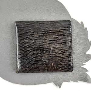 May include: A dark brown bifold wallet featuring a textured, reptilian-inspired scale pattern across its surface. The wallet has a rectangular shape with clean, straight edges and a glossy finish that highlights the intricate, small-scale grid design.