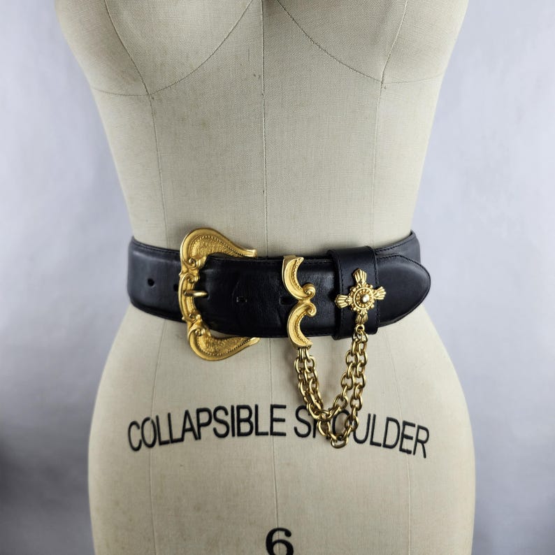 Puede incluir: Cintur&oacute;n de cuero negro con una hebilla dorada grande y detalles dorados decorativos. El cintur&oacute;n presenta una cadena dorada y un adorno en forma de cruz. Se muestra en un maniqu&iacute;.