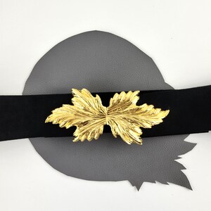 May include: A black belt with a gold-colored leaf-shaped buckle. The buckle is the focal point, with intricate details and a shiny finish. The belt is set against a gray background.