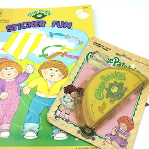 Może przedstawiać: Książka do kolorowania Cabbage Patch Kids "Sticker Fun" z żółtą okładką i obrazkiem latawca. Książka leży obok torebki z lusterkiem i grzebieniem Cabbage Patch Kids w żółto-zielonym wzorze.