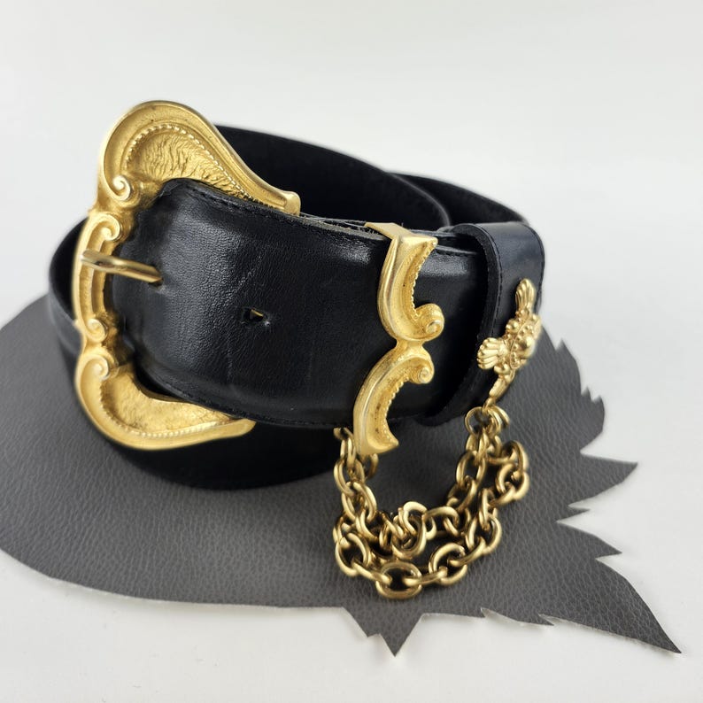 Puede incluir: Cintur&oacute;n de cuero negro con una hebilla dorada grande y ornamentada y detalles dorados decorativos. Una cadena dorada cuelga del cintur&oacute;n. El cintur&oacute;n est&aacute; sobre una superficie gris.