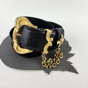 Puede incluir: Un cintur&oacute;n de cuero negro con una hebilla dorada grande y ornamentada. El cintur&oacute;n presenta elementos decorativos dorados adicionales y una cadena colgante. Probablemente sea un accesorio de moda vintage.
