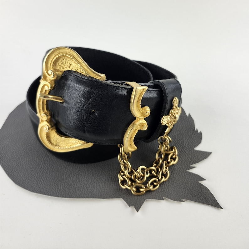 Puede incluir: Un cintur&oacute;n de cuero negro con una hebilla dorada grande y ornamentada. El cintur&oacute;n presenta elementos decorativos dorados adicionales y una cadena colgante. Probablemente sea un accesorio de moda vintage.