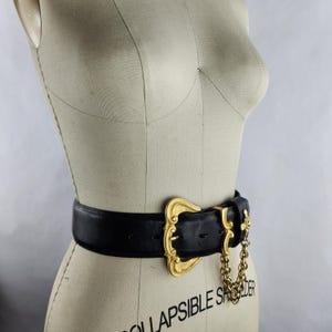 Puede incluir: Cintur&oacute;n de cuero negro con una hebilla dorada grande y ornamentada. La hebilla presenta un dise&ntilde;o intrincado y est&aacute; conectada a una cadena dorada. El cintur&oacute;n se muestra en un maniqu&iacute;.