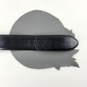 Puede incluir: Un cintur&oacute;n de cuero negro con un extremo redondeado y varios agujeros para ajustar. El cintur&oacute;n est&aacute; colocado sobre un recorte de cuero gris en forma de hoja, sobre un fondo blanco. La textura y el dise&ntilde;o del cintur&oacute;n son claramente visibles.