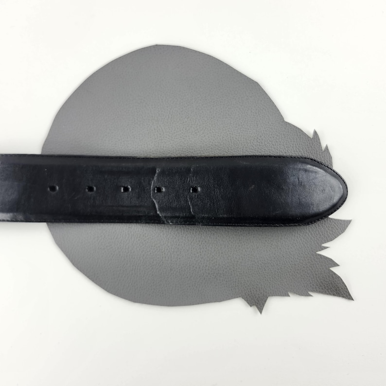 Puede incluir: Un cintur&oacute;n de cuero negro con un extremo redondeado y varios agujeros para ajustar. El cintur&oacute;n est&aacute; colocado sobre un recorte de cuero gris en forma de hoja, sobre un fondo blanco. La textura y el dise&ntilde;o del cintur&oacute;n son claramente visibles.