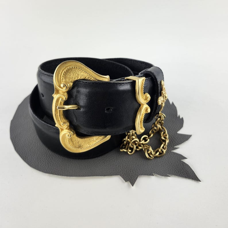 Puede incluir: Cintur&oacute;n de cuero negro con una hebilla dorada grande y ornamentada y detalles de cadena dorada a juego. La hebilla presenta un dise&ntilde;o decorativo. El cintur&oacute;n est&aacute; enrollado sobre una superficie gris en forma de hoja.