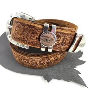 May include: A brown leather belt featuring an embossed floral pattern and silver-toned metal hardware. The buckle and a sliding metal loop are engraved with the text Alien Workshop and feature a small winged alien head logo design.