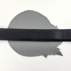 Puede incluir: Un cintur&oacute;n de cuero negro se coloca sobre un trozo de cuero gris de forma irregular. El cintur&oacute;n est&aacute; centrado y parece tener un ancho est&aacute;ndar. El fondo es blanco.