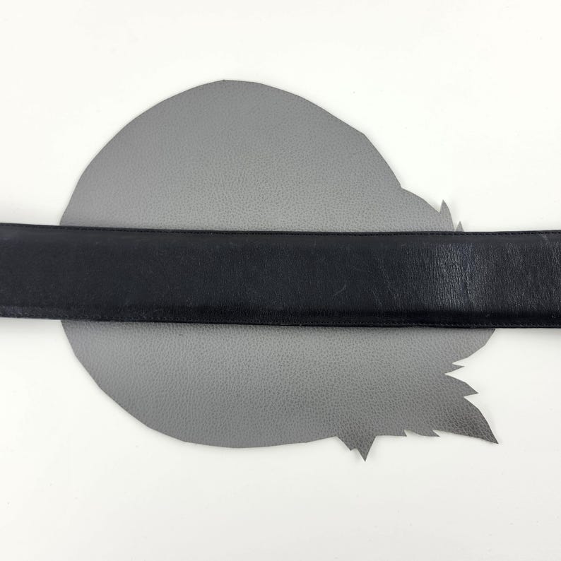 Puede incluir: Un cintur&oacute;n de cuero negro se coloca sobre un trozo de cuero gris de forma irregular. El cintur&oacute;n est&aacute; centrado y parece tener un ancho est&aacute;ndar. El fondo es blanco.