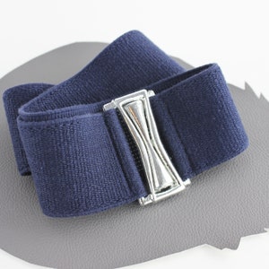 Könnte beinhalten: Ein marineblauer elastischer Gürtel mit einem silberfarbenen Metallverschluss. Der Gürtel besteht aus einem weichen, strukturierten Stoff und ist für das Tragen um die Taille konzipiert. Der Verschluss ist ein dekoratives Element.
