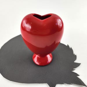 May include: A glossy red heart-shaped vase. The vase has a rounded body and a small, circular base. The top of the vase has a heart-shaped opening. The vase is sitting on a dark gray, leaf-shaped surface.