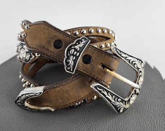 Puede incluir: Un cinturón de cuero marrón con detalles metálicos plateados. El cinturón presenta una hebilla decorativa, detalles de tachuelas y adornos plateados. El cinturón es adecuado para uso casual o de estilo western.