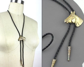May include: A black bolo tie with a gold horse head pendant. The tie is on a mannequin and the pendant is facing the viewer.