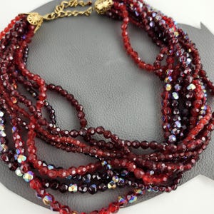 Puede incluir: Un collar multi-cadena hecho de cuentas de vidrio rojo e iridiscente. El collar está hecho de varias cadenas de cuentas, y las cuentas son de diferentes tamaños y formas. El collar es una pieza de joyería hermosa y elegante.