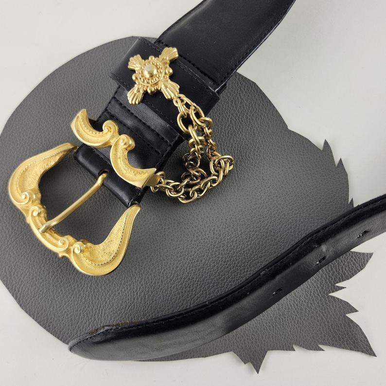 Puede incluir: Cintur&oacute;n de cuero negro con hebilla dorada y detalles decorativos. El cintur&oacute;n presenta una cadena dorada y un adorno en forma de cruz. La hebilla tiene un dise&ntilde;o ornamentado y curvado.