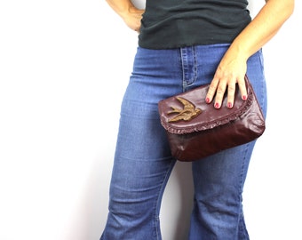 May include: A maroon leather clutch with a gold bird embellishment. The clutch has a ruffled edge and is held by a person wearing blue jeans.