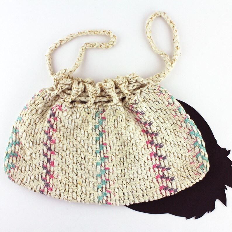 May include: A white crocheted bag with pink and blue stripes. The bag has a drawstring closure and a long strap.