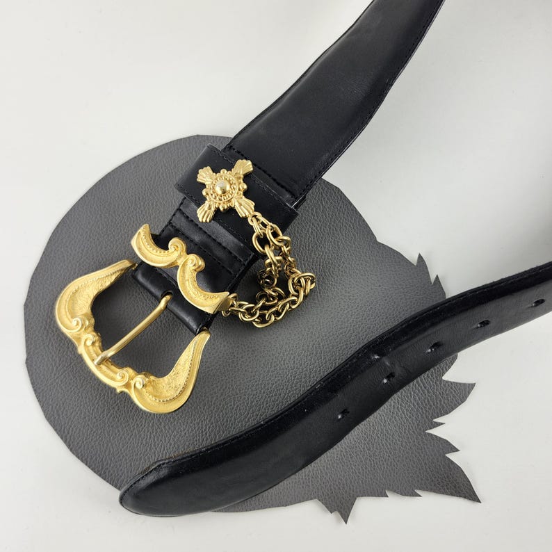 Puede incluir: Cintur&oacute;n de cuero negro con una hebilla dorada ornamentada y una cadena decorativa. La hebilla tiene un dise&ntilde;o detallado y el cintur&oacute;n est&aacute; sobre un fondo gris. Un accesorio cl&aacute;sico.