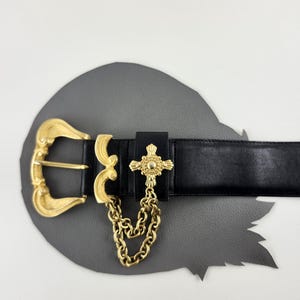 Puede incluir: Cintur&oacute;n de cuero negro con hebilla dorada y un colgante decorativo de cruz. El cintur&oacute;n presenta una cadena dorada y un dise&ntilde;o de hebilla detallado. El cintur&oacute;n se muestra sobre una superficie gris.