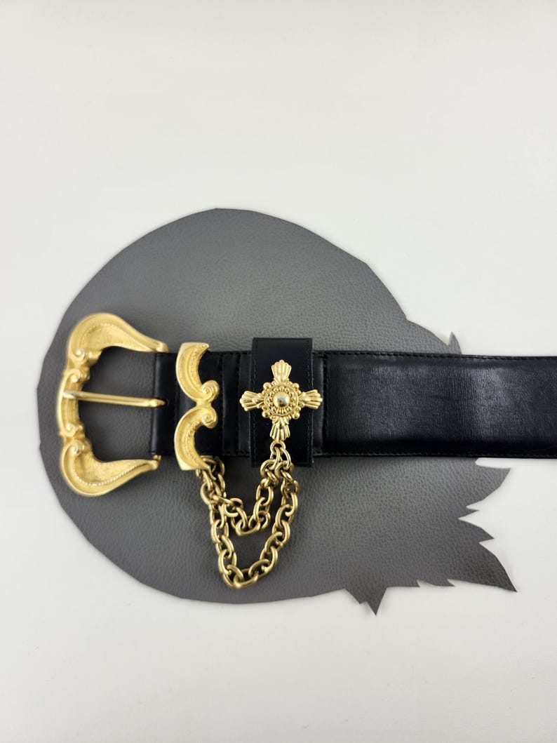 Puede incluir: Cintur&oacute;n de cuero negro con hebilla dorada y un colgante decorativo de cruz. El cintur&oacute;n presenta una cadena dorada y un dise&ntilde;o de hebilla detallado. El cintur&oacute;n se muestra sobre una superficie gris.