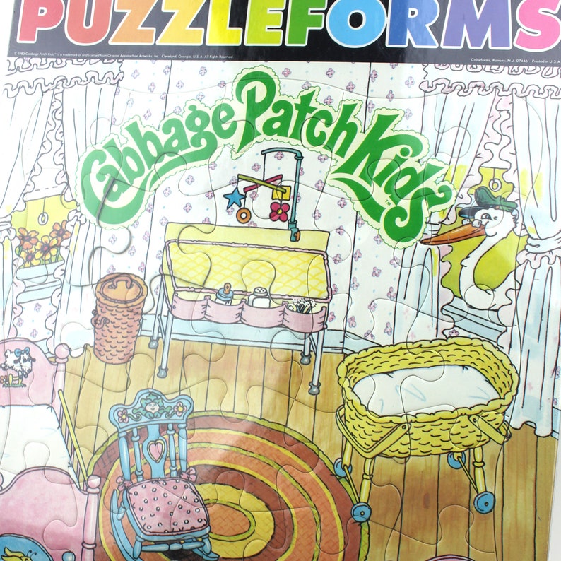 Puede incluir: Un rompecabezas vintage de Cabbage Patch Kids que presenta una escena de guarder&iacute;a con una cuna, una mecedora, un cambiador y una cig&uuml;e&ntilde;a mirando por una ventana. Las piezas del rompecabezas tienen forma de rompecabezas y el fondo es un piso de madera con una alfombra.