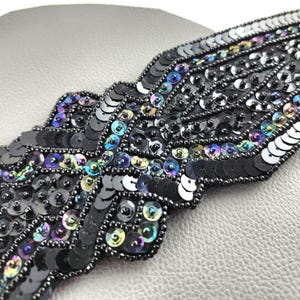 May include: Close-up of a black sequin applique with iridescent accents. The applique is in a wing or feather shape, with rows of black sequins and small, dark beads. The sequins catch the light, creating a shimmering effect.