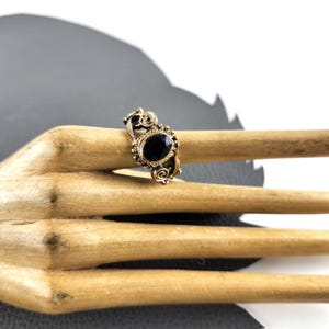 Könnte beinhalten: Ein Ring im Vintage-Stil mit einem ovalen, schwarzen Edelstein, der in einem goldfarbenen Metall gefasst ist. Der Ring hat kunstvolle Schnörkel und eine detaillierte Lünettenfassung. Der Ring wird auf einer Holzoberfläche präsentiert.