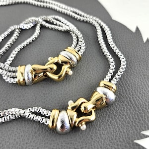 Può includere: Primo piano di una collana argentata con chiusure dorate. La collana presenta un design a catena con una costruzione a tre fili. Le chiusure hanno un design decorativo con un piccolo accento di perla.