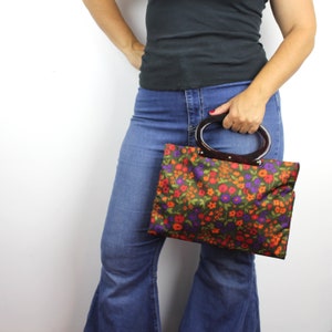 Op de afbeelding: Een handtas met bloemenprint en een bruine kunststof handvat. De tas wordt vastgehouden door iemand die een blauwe jeans draagt.
