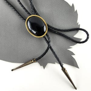 May include: A bolo tie featuring a large, oval black stone set in a gold-toned metal frame. The braided black leather cord has decorative metal tips. The tie is displayed on a gray textured surface.