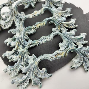 Op de afbeelding: Vier sierlijke, decoratieve ladegrepen met een verweerde, wit-groene verfbehandeling. De grepen hebben een rolvormig ontwerp met ingewikkelde details.