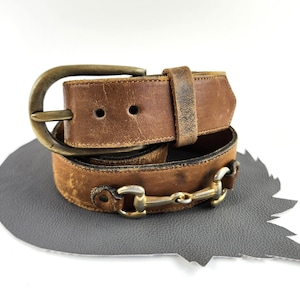 May include: A brown leather belt featuring a distressed finish, a rounded brass-toned buckle, and a metallic horse bit detail integrated into the strap. The belt is coiled, showing visible wear and texture on the leather surface and metal hardware.