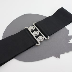 Könnte beinhalten: Ein schwarzer Gummibandgürtel mit einer silbernen Schnalle mit drei runden Metallakzenten. Der Gürtel liegt auf einer grauen Oberfläche.