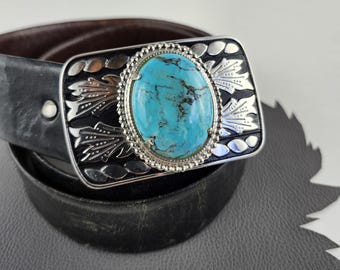 May include: A rectangular silver belt buckle with a large oval turquoise stone at the center. The buckle has a black background with silver decorative elements and is attached to a black leather belt.