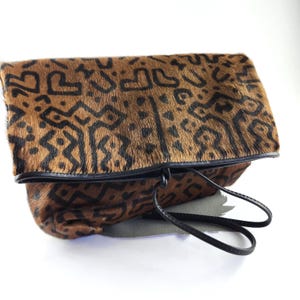 May include: A brown and black patterned clutch bag made of textured hair-on hide. The rectangular bag features a fold-over flap with a repeating geometric design in black. It includes a thin black leather wrist strap and black piping edges.