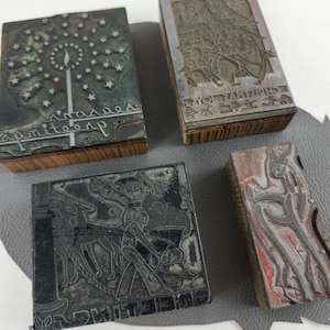 May include: Four rectangular letterpress printing blocks with wooden bases and metal relief surfaces. One block features a candle surrounded by stars, another shows a stylized animal, and two depict intricate scenes with figures and text.