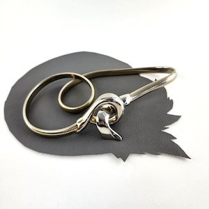 Könnte beinhalten: Ein flexibler Gürtel aus goldfarbener Metall-Schlangenkette mit einer polierten, silberfarbenen Schnalle in Knotenform. Der Gürtel ist kreisförmig aufgerollt und zeigt die gerippte Textur des Metallbandes sowie die glatte, reflektierende Oberfläche.