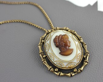 May include: A gold chain necklace with a cameo pendant. The cameo is oval shaped and features a brown profile of a woman with a white background. The pendant is surrounded by a gold frame with a decorative design.