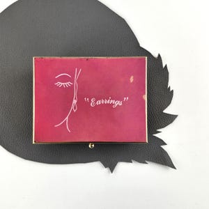 May include: A vintage jewelry box with a gold frame and a red lid. The lid features a minimalist face illustration and the word "Earrings" in white script. The box is set on a black leather-like surface.