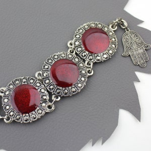 Puede incluir: Una pulsera de plata con tres abalorios redondos de esmalte rojo y un abalorio hamsa de plata. Los abalorios están decorados con detalles intrincados.